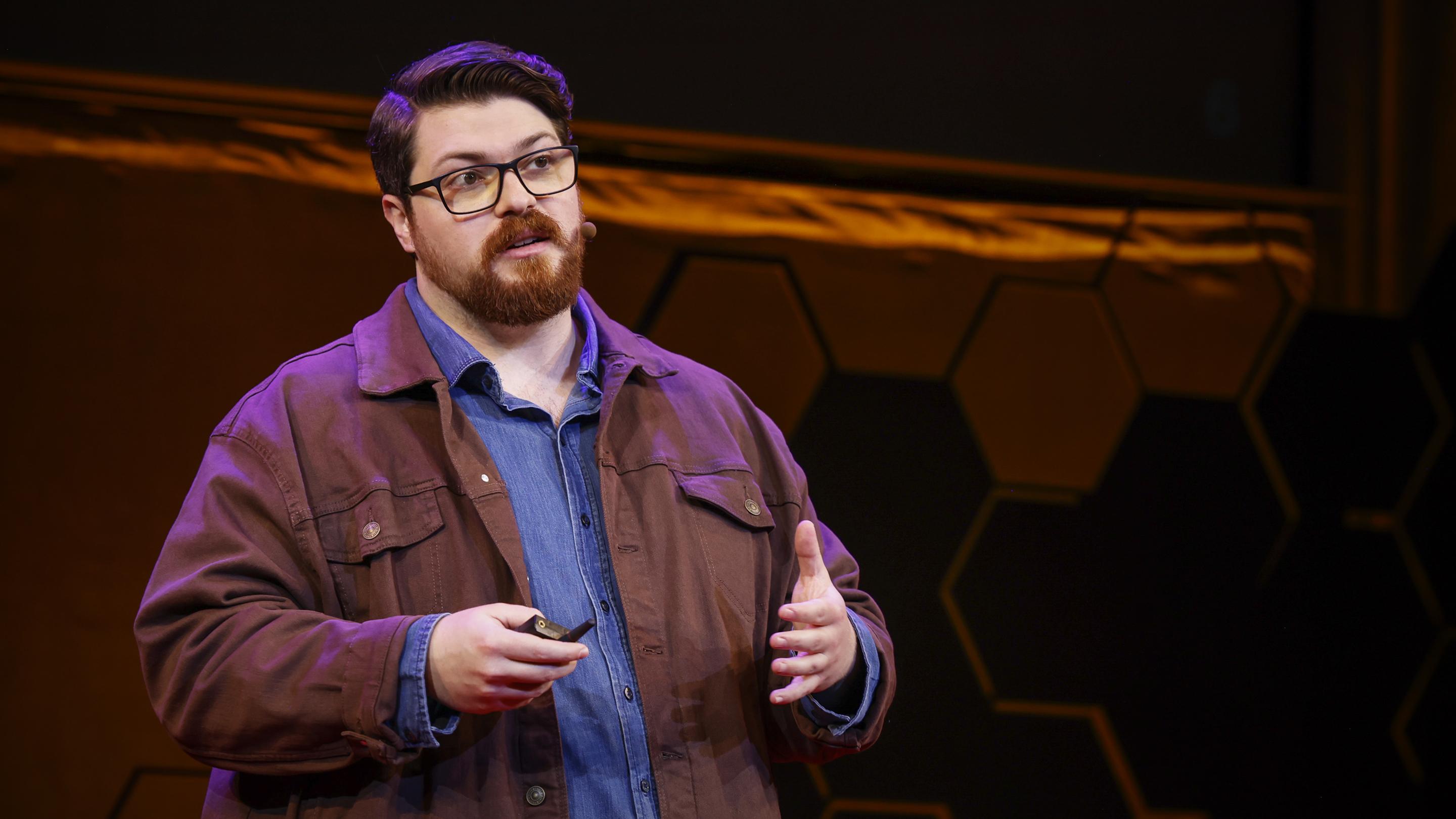 portrait of Guillaume Verdon speaking at TED AI 2024, captured mid-presentation on stage with a confident expression, highlighting his role as a thought leader in advancing energy-efficient AI through thermodynamic computing and quantum innovations.